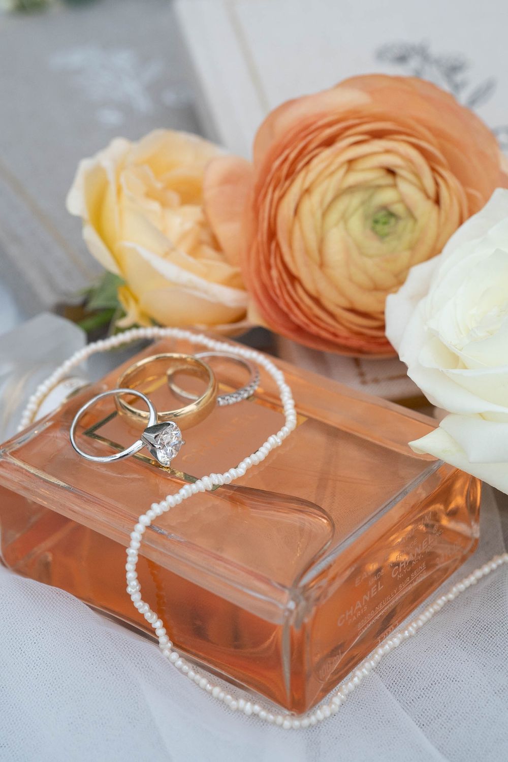Diamond engagement ring and pearl necklace displayed on peach-colored surface with ranunculus flowers.