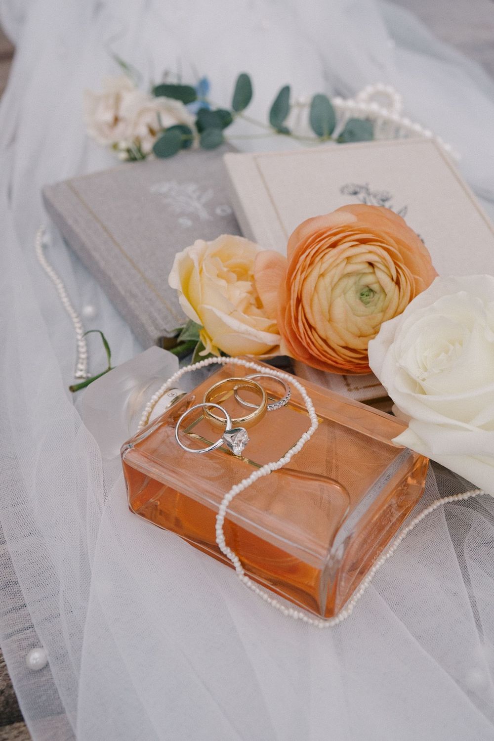 Elegant wedding details with perfume bottle, roses, and invitation books arranged on delicate tulle fabric.