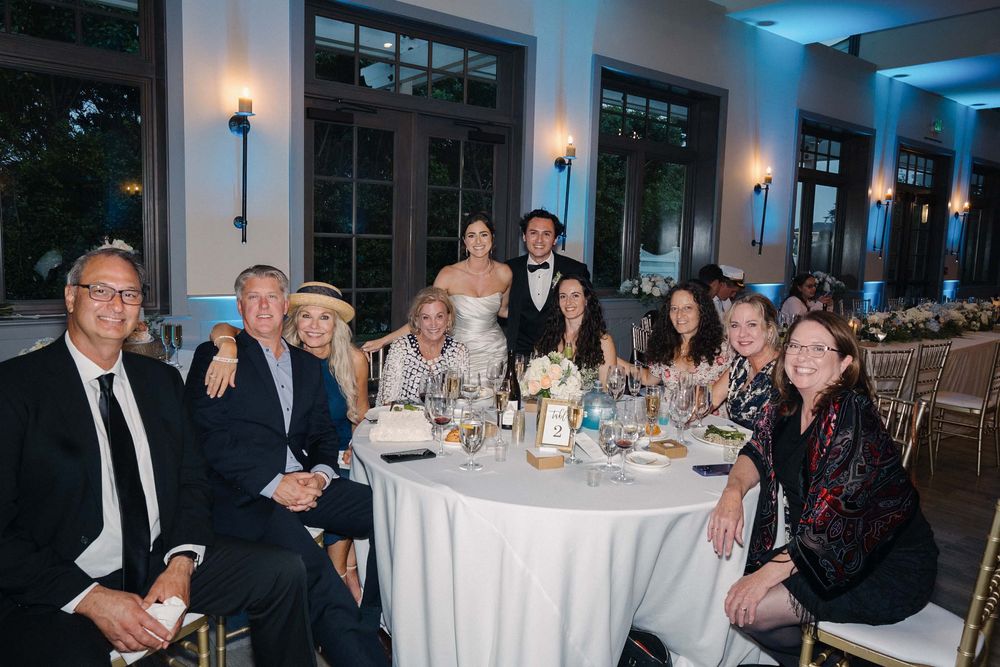 A group of formally dressed guests seated at a round table during an elegant evening reception in a well-lit venue.