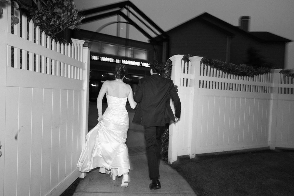 A black and white wedding photo shows a couple walking away from the camera on a path beside a white fence.