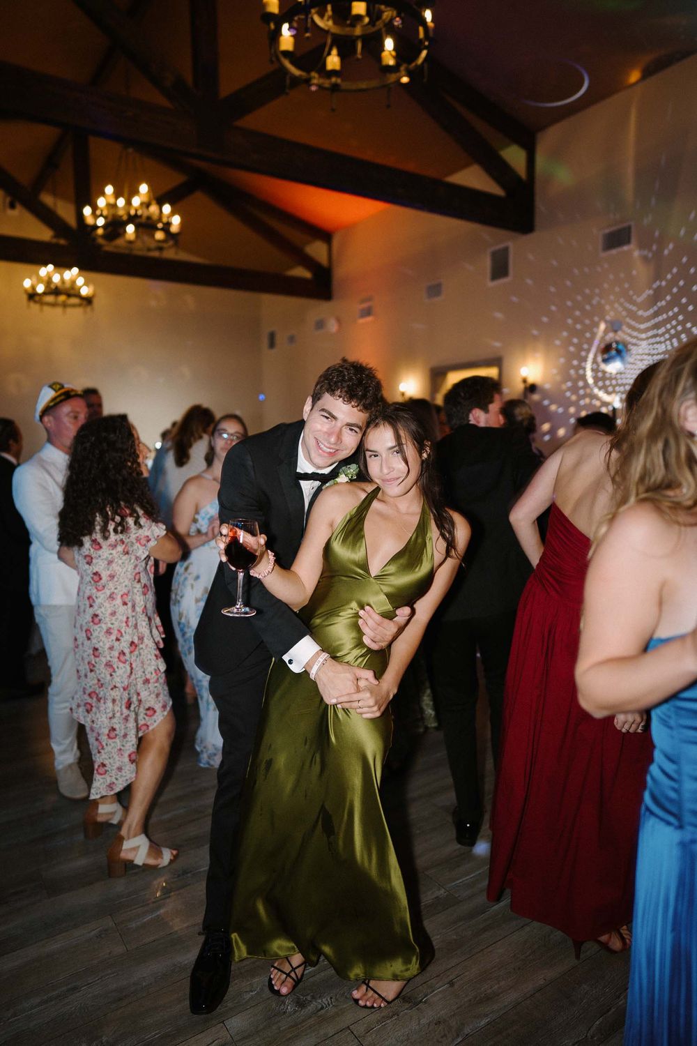 Wedding guests dancing together on a dark wooden dance floor under warm lighting and chandeliers at a reception venue.