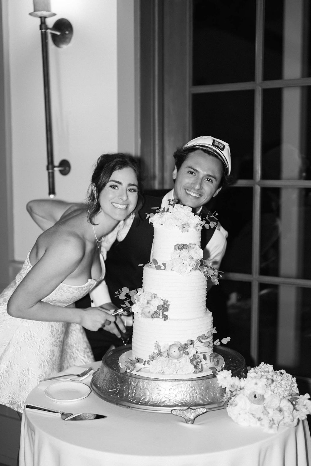 A black and white photo of a couple posing with their elegant white wedding cake decorated with flowers.