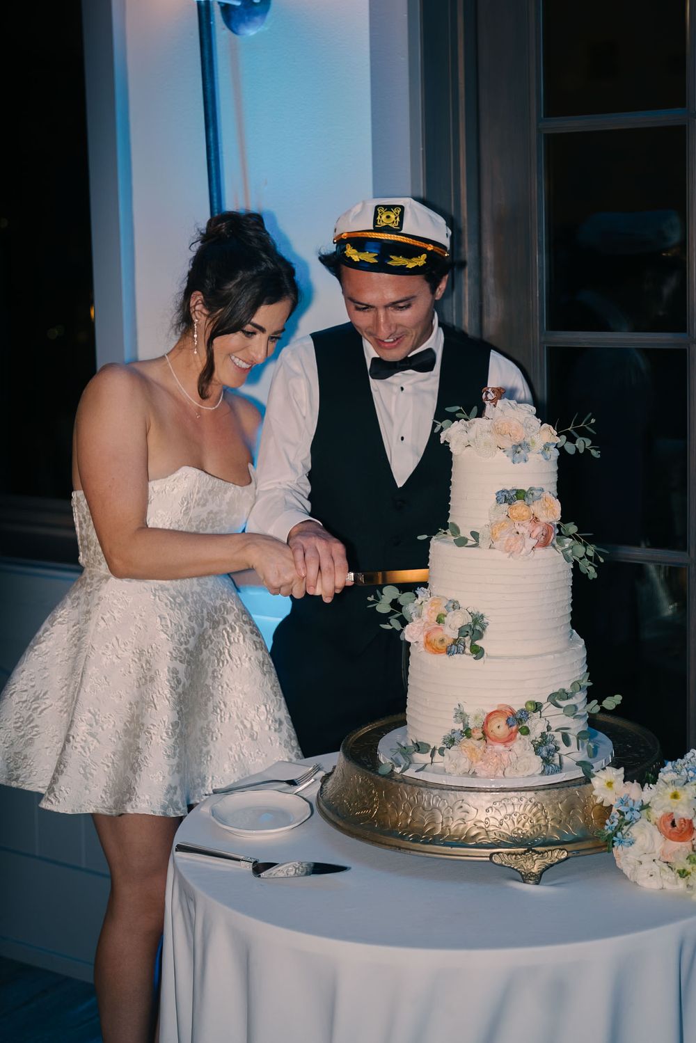 A couple cuts their elegant three-tiered wedding cake decorated with peach and white flowers on a silver stand.