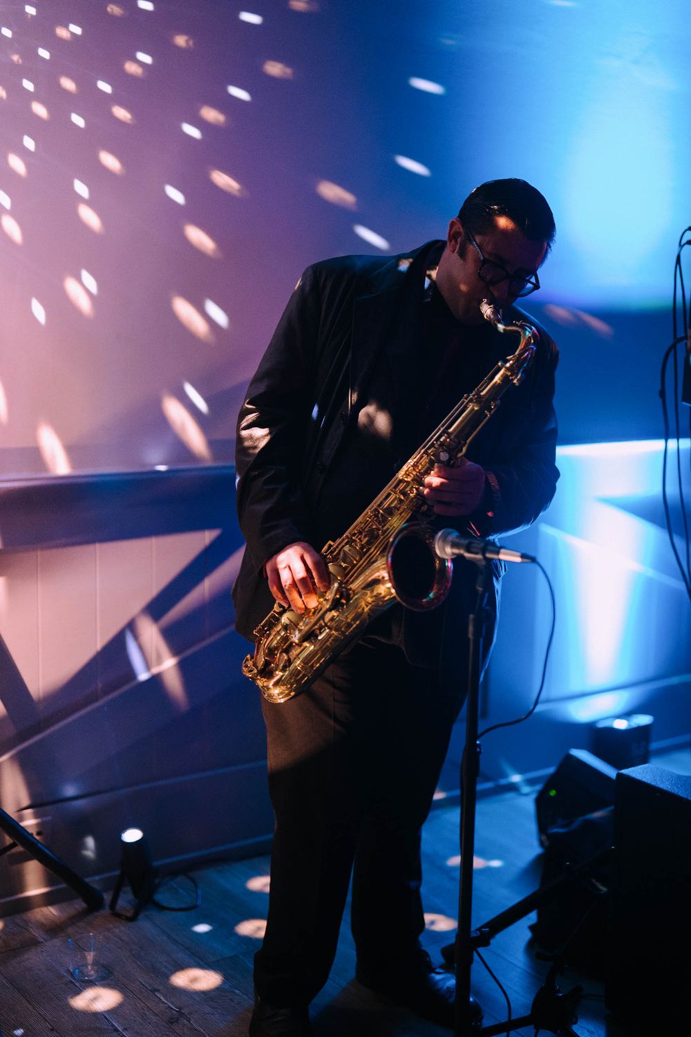 Silhouetted saxophone player performs on stage with dramatic blue lighting and spotlights creating scattered light effects.
