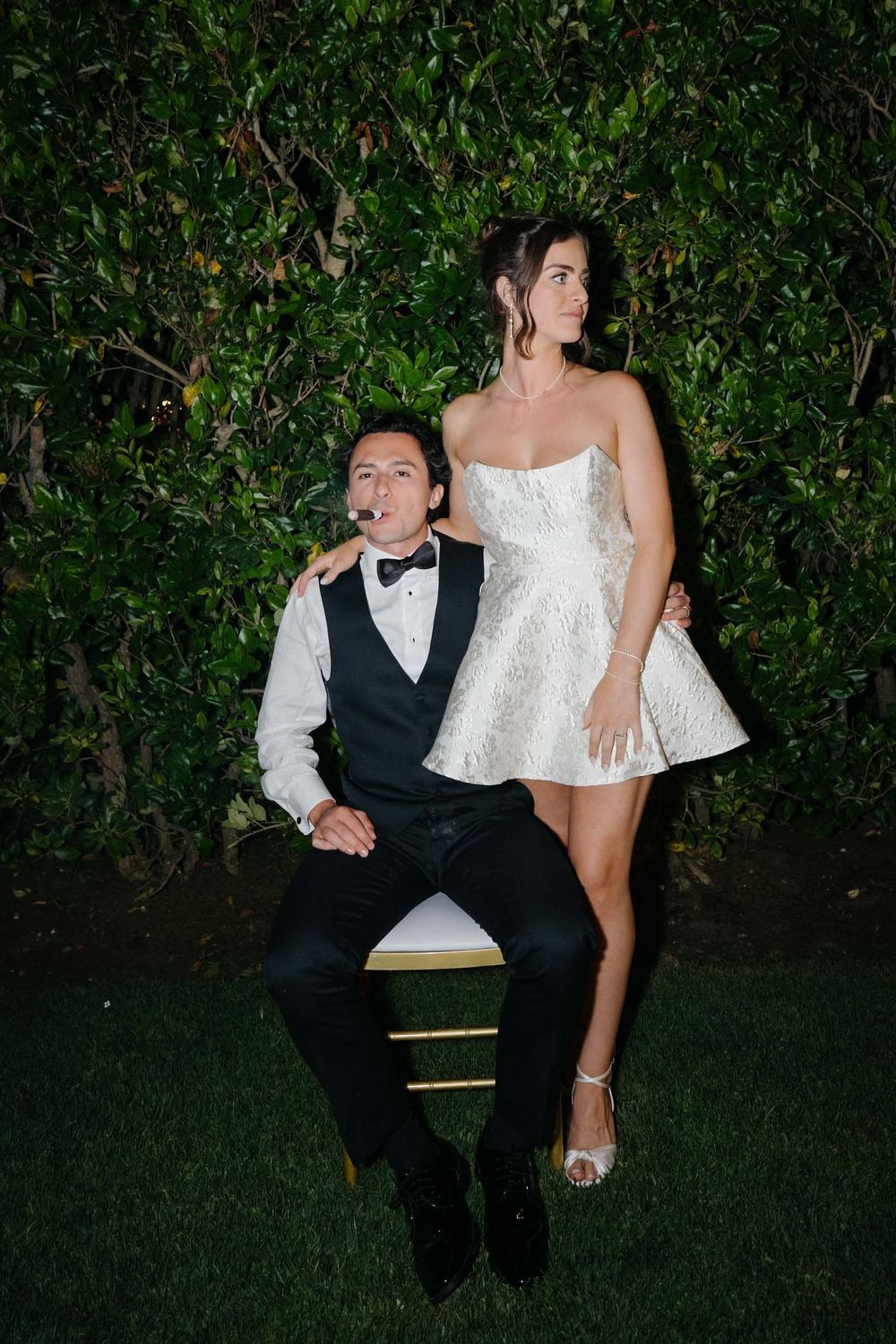 A couple poses playfully against a hedge wall backdrop, with one sitting and one standing in formal attire.