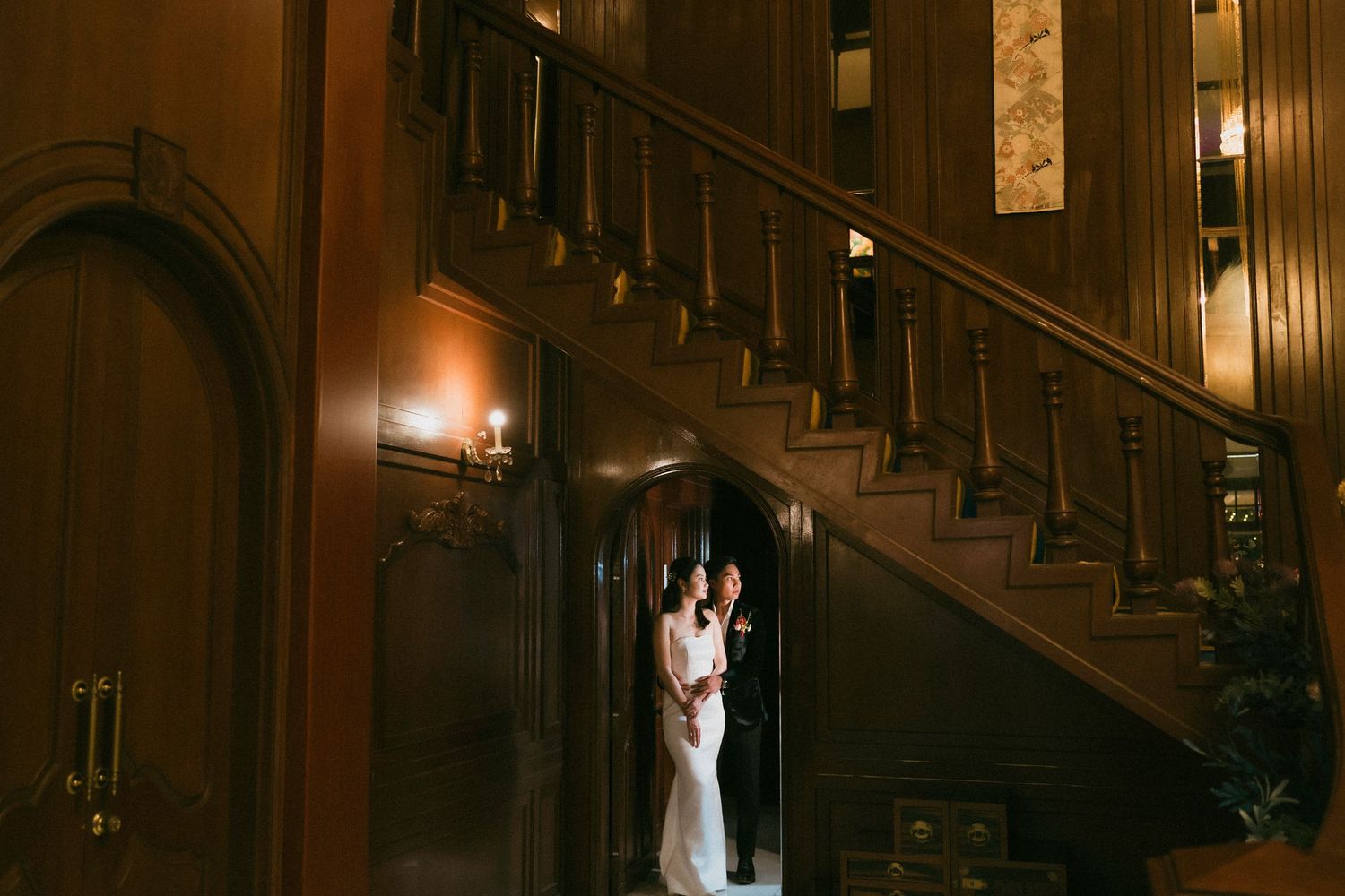 Couple embracing in a warmly lit wooden staircase alcove inside an elegant historic mansion.