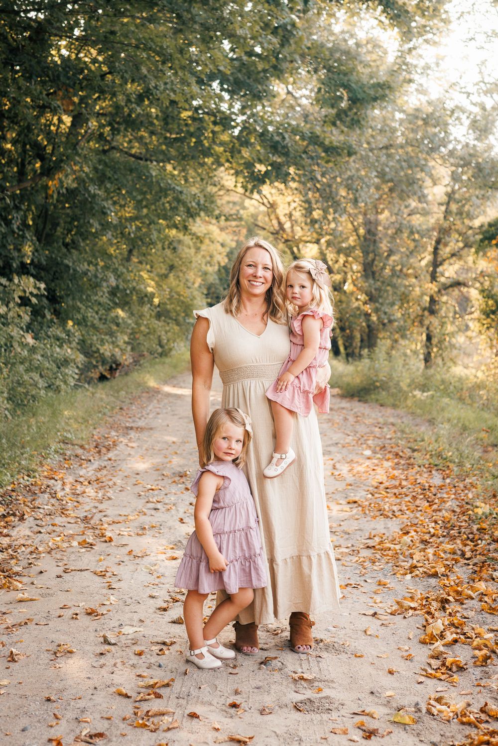 A family wearing light colored dresses poses together on a fall path covered in orange and yellow leaves at sunset.