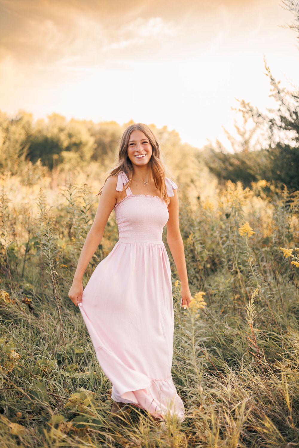 A person in a flowing pink dress stands in a sunlit field of tall grass during golden hour.