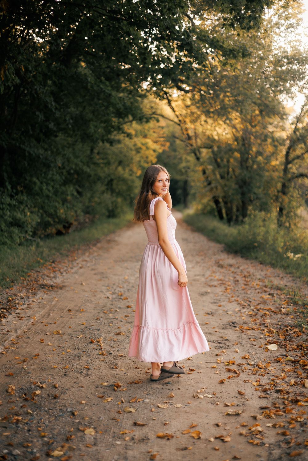 A figure in a flowing pink dress walks down a leaf-covered autumn path surrounded by golden trees at sunset.