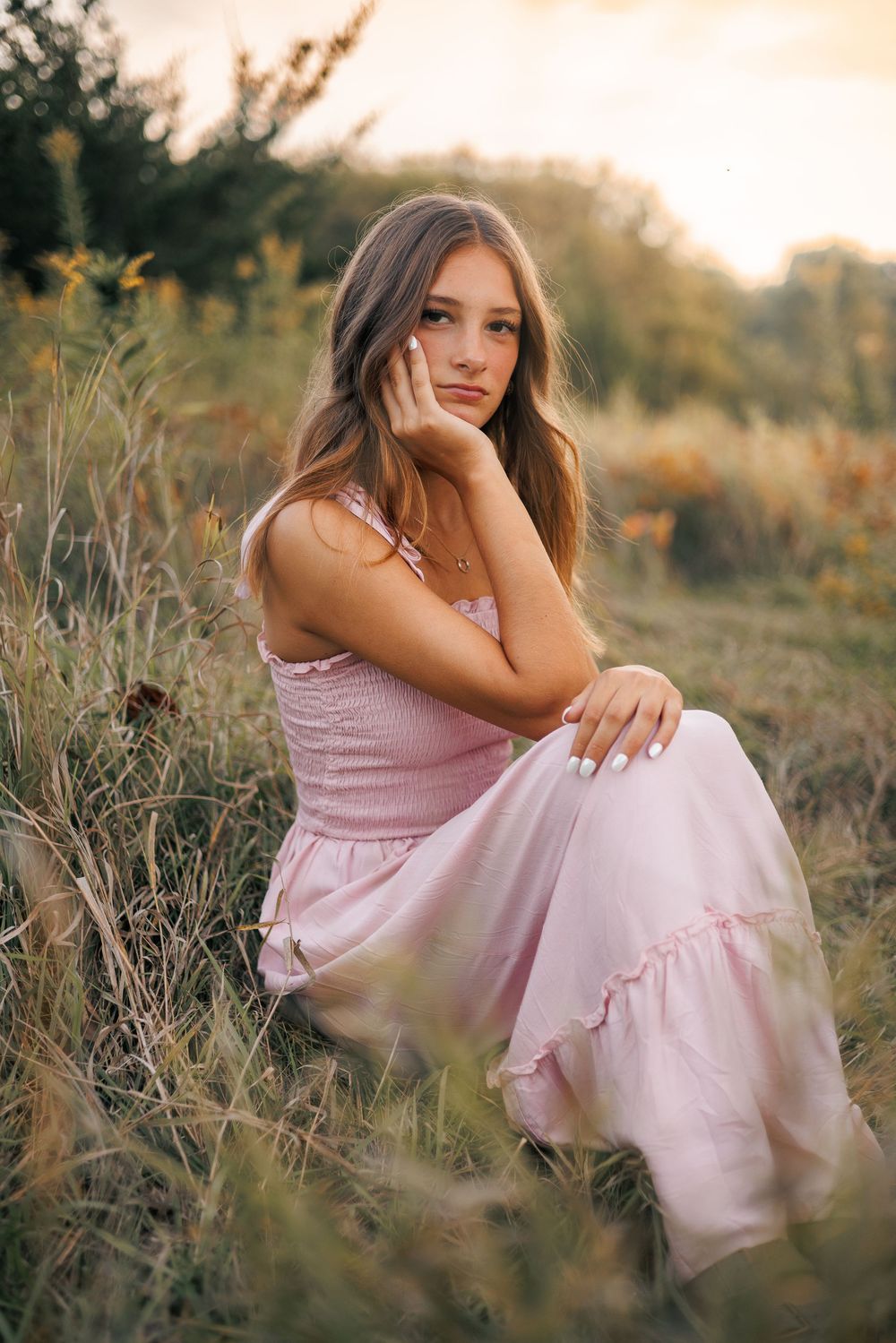 Ethereal outdoor portrait series in soft pink dress sitting among wild grass during golden hour sunset.