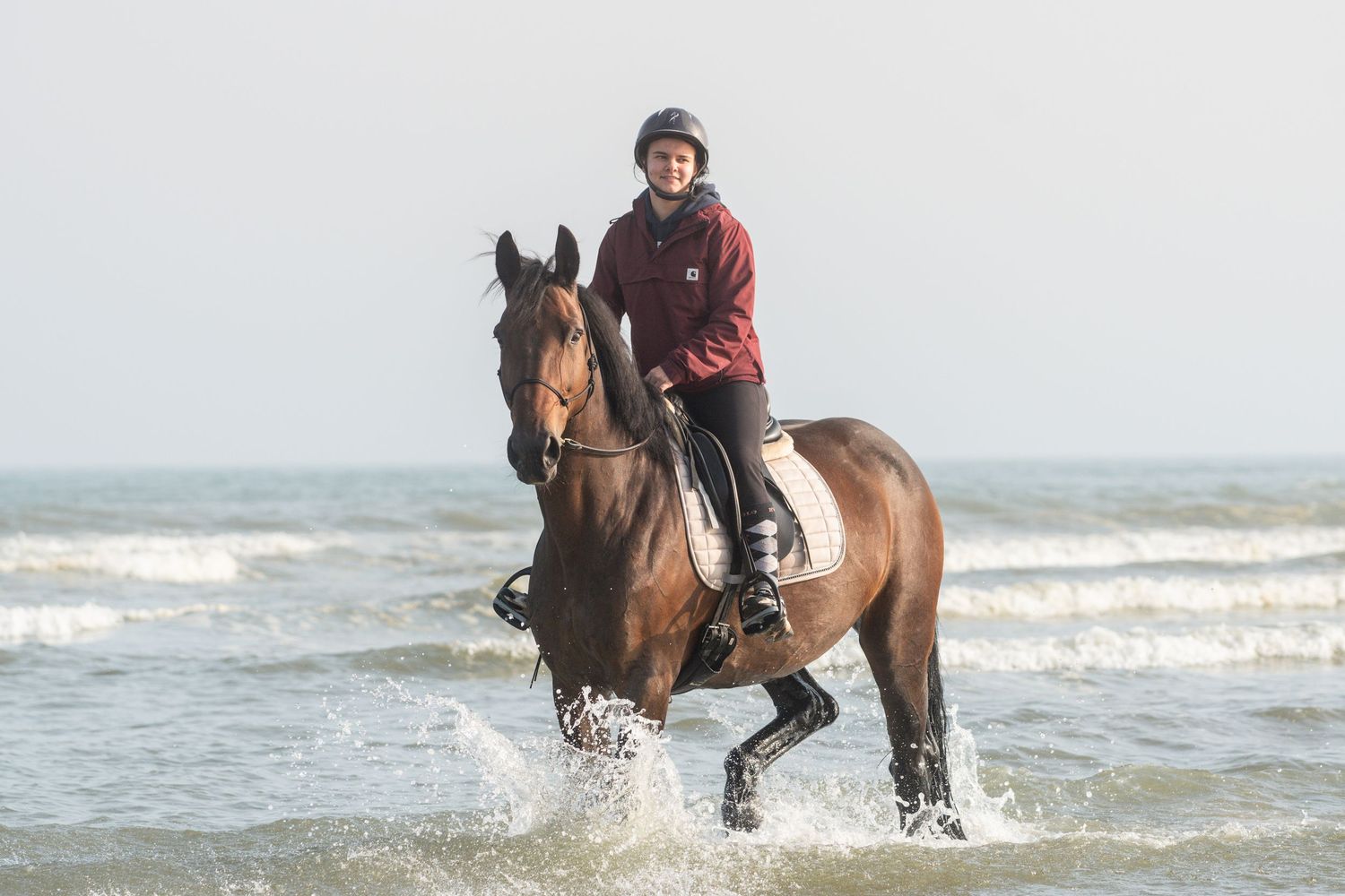 Horse and rider splashing through waves at the beach during cloudy day.