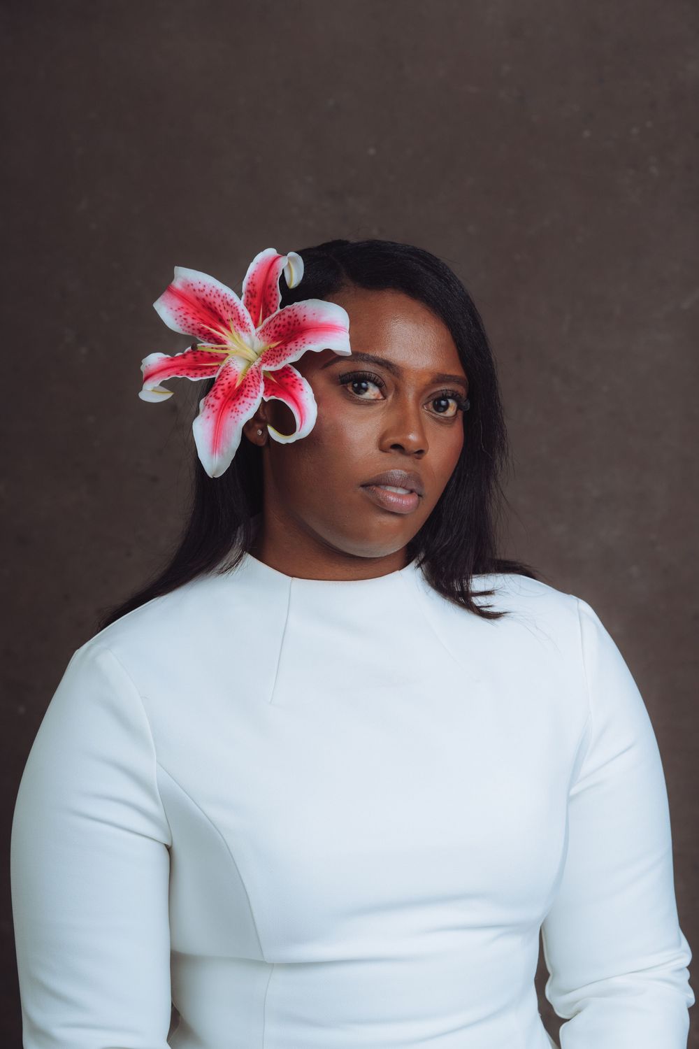 Portrait of a person in white wearing a pink lily flower in their hair against a dark background.
