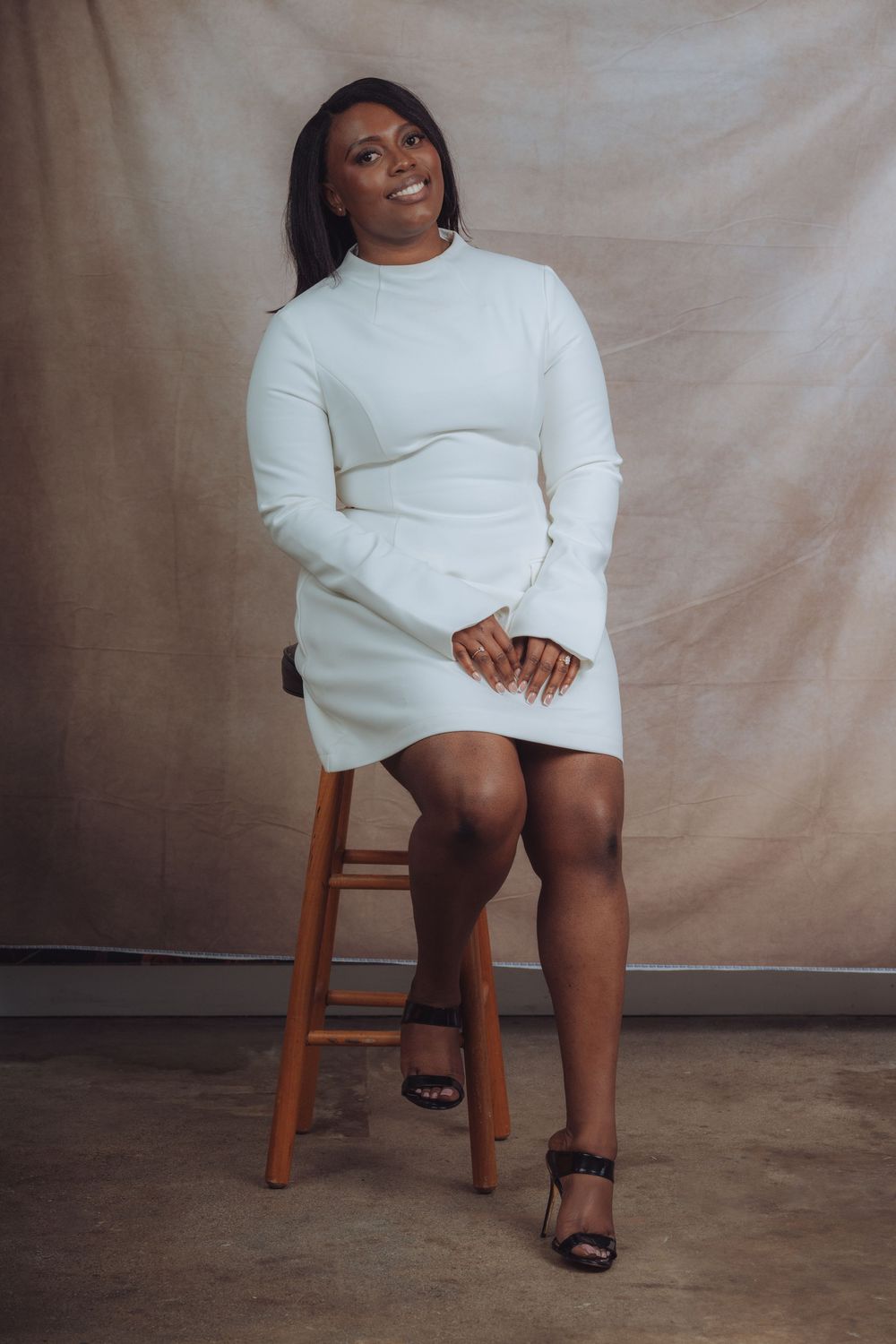 Minimalist fashion shoot on wooden stool showcasing white sweater dress and elegant footwear.