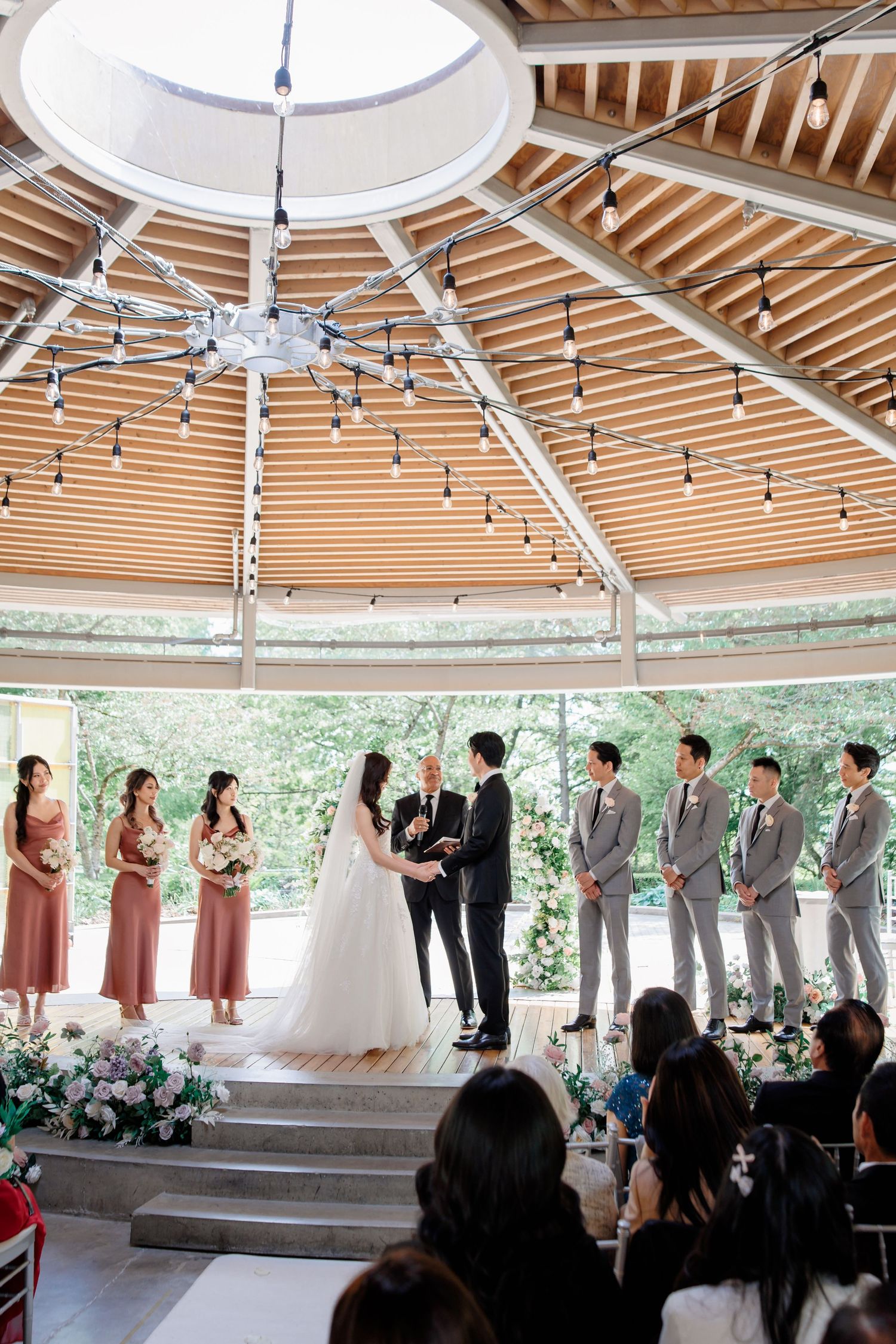 Wedding ceremony taking place in an elegant gazebo with wooden beams and skylight under natural lighting.