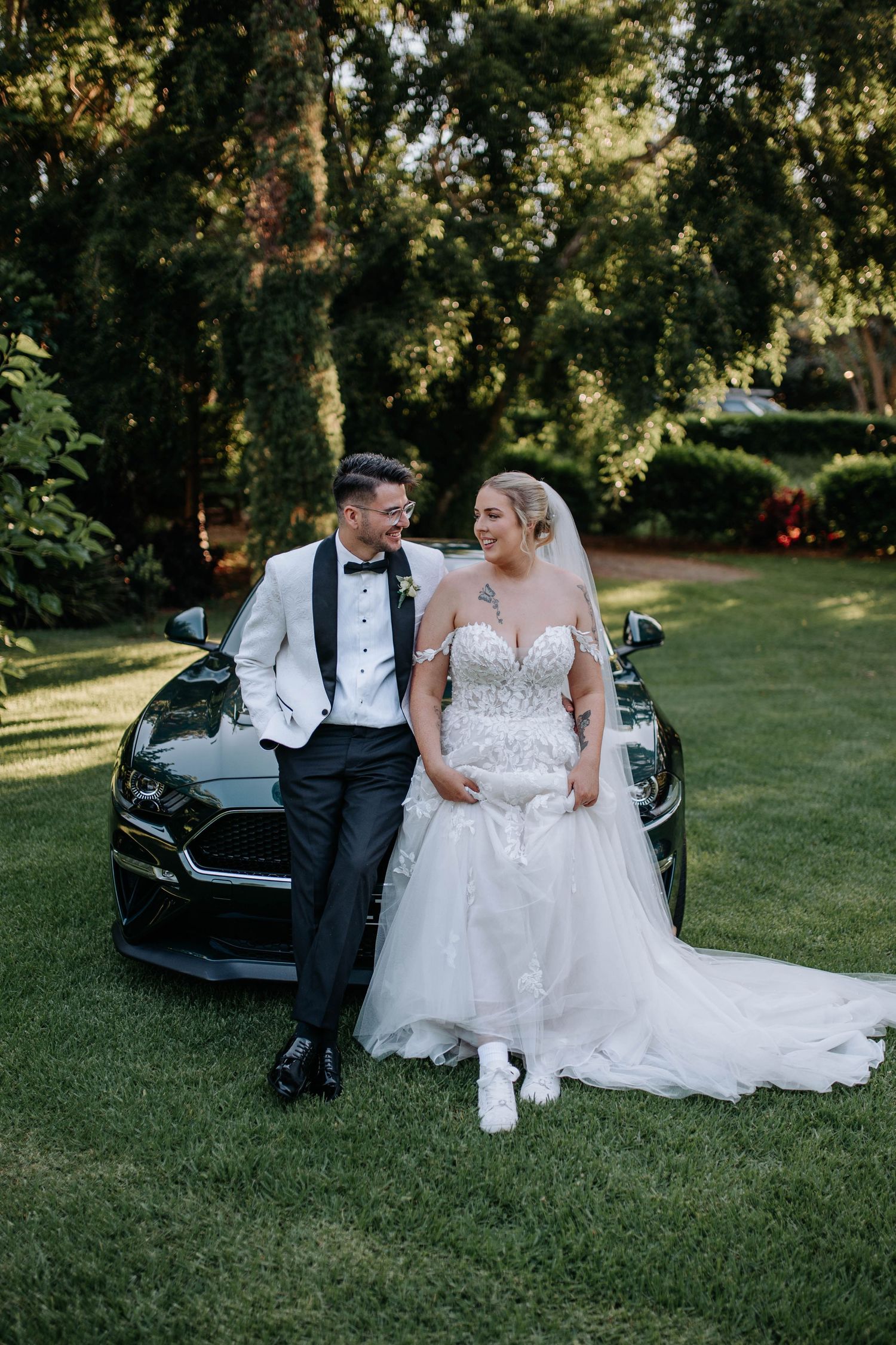 A couple in wedding attire sits together on grass near a black luxury car during their outdoor photo session.