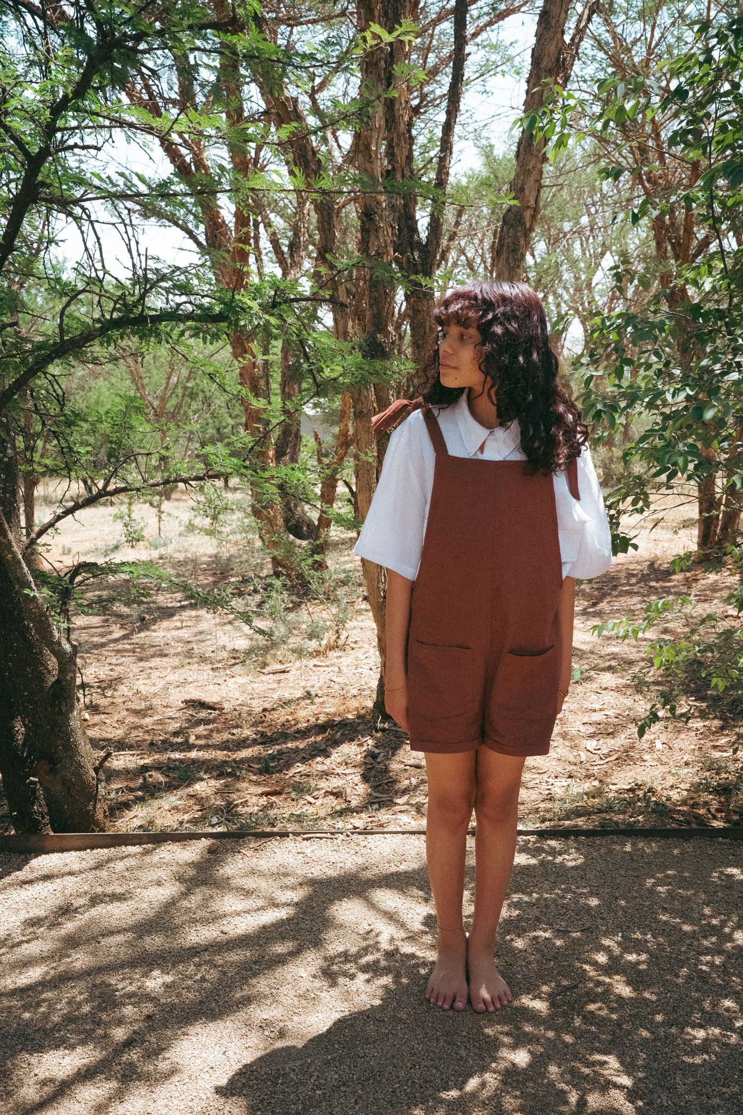 Person in brown overalls standing among trees in a sunlit wooded area.