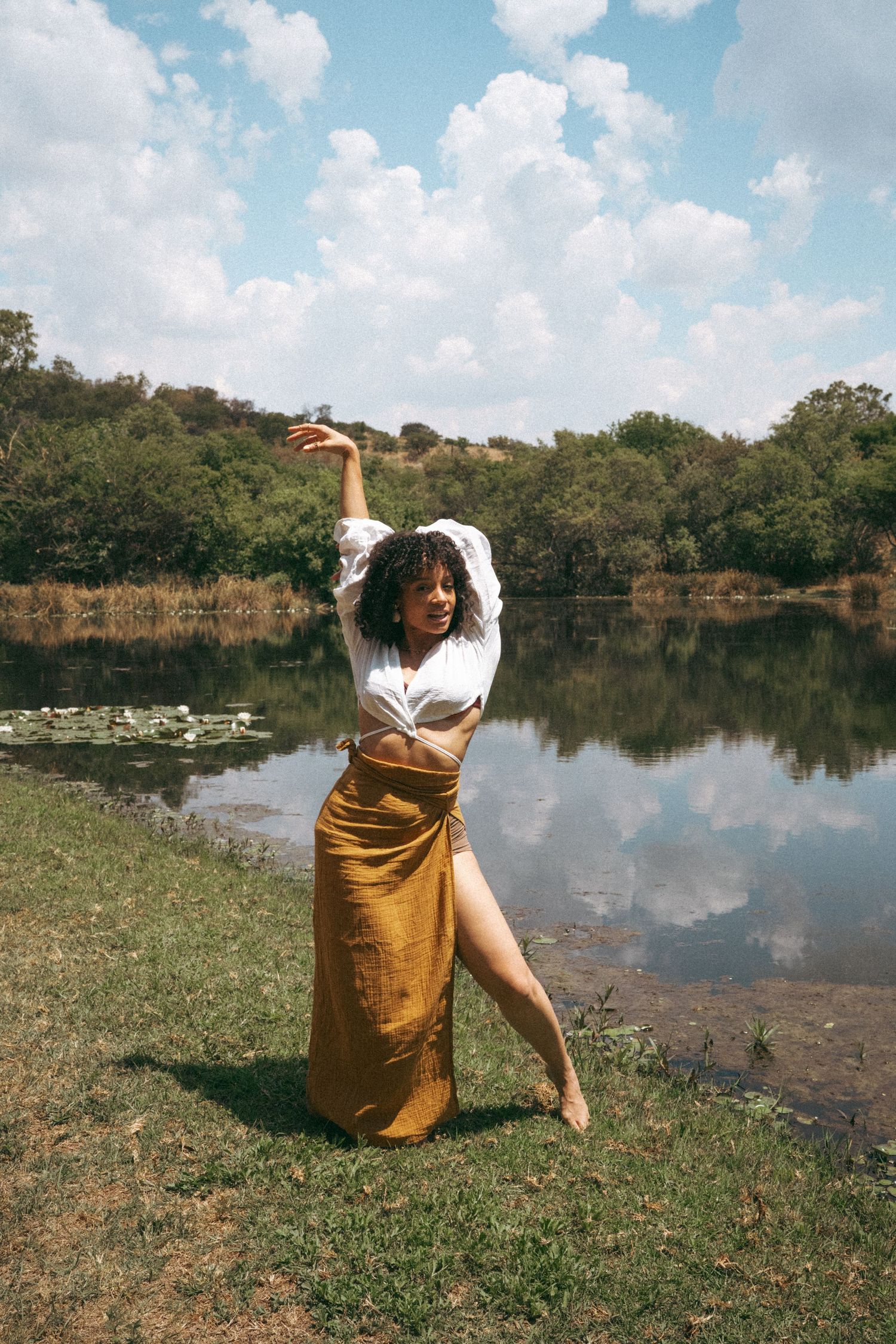 Artistic outdoor portraits by a lake featuring white top and mustard skirt against blue sky.