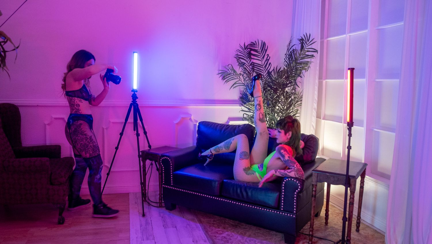Behind-the-scenes photo shoot with photographer and model on blue velvet couch in pink-lit studio space.