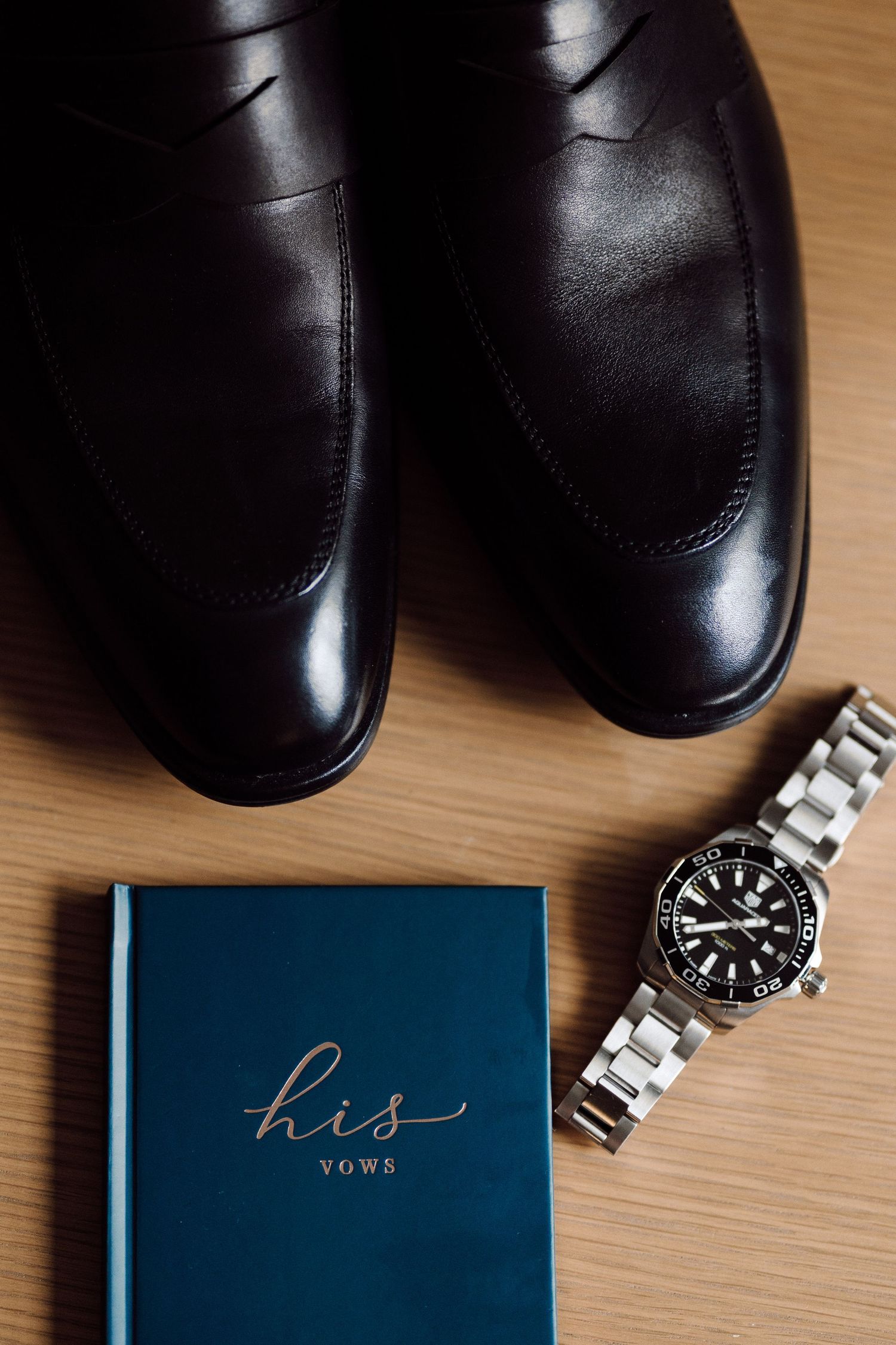 Black dress shoes and a silver watch arranged next to a blue book labeled 'his' on wooden surface.