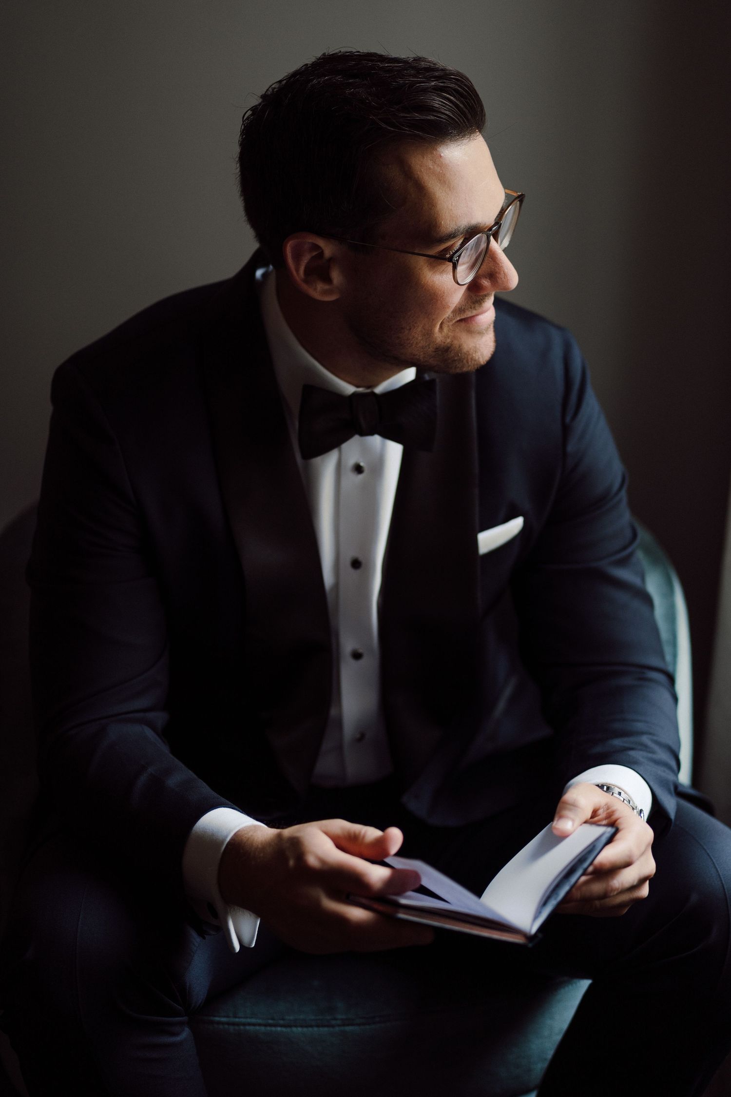 Moody portraits of groom in tuxedo reading while seated in dramatic lighting.