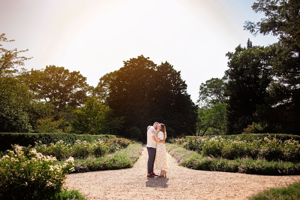 A romantic couple stands together on a scenic gravel path surrounded by lush greenery at sunset.