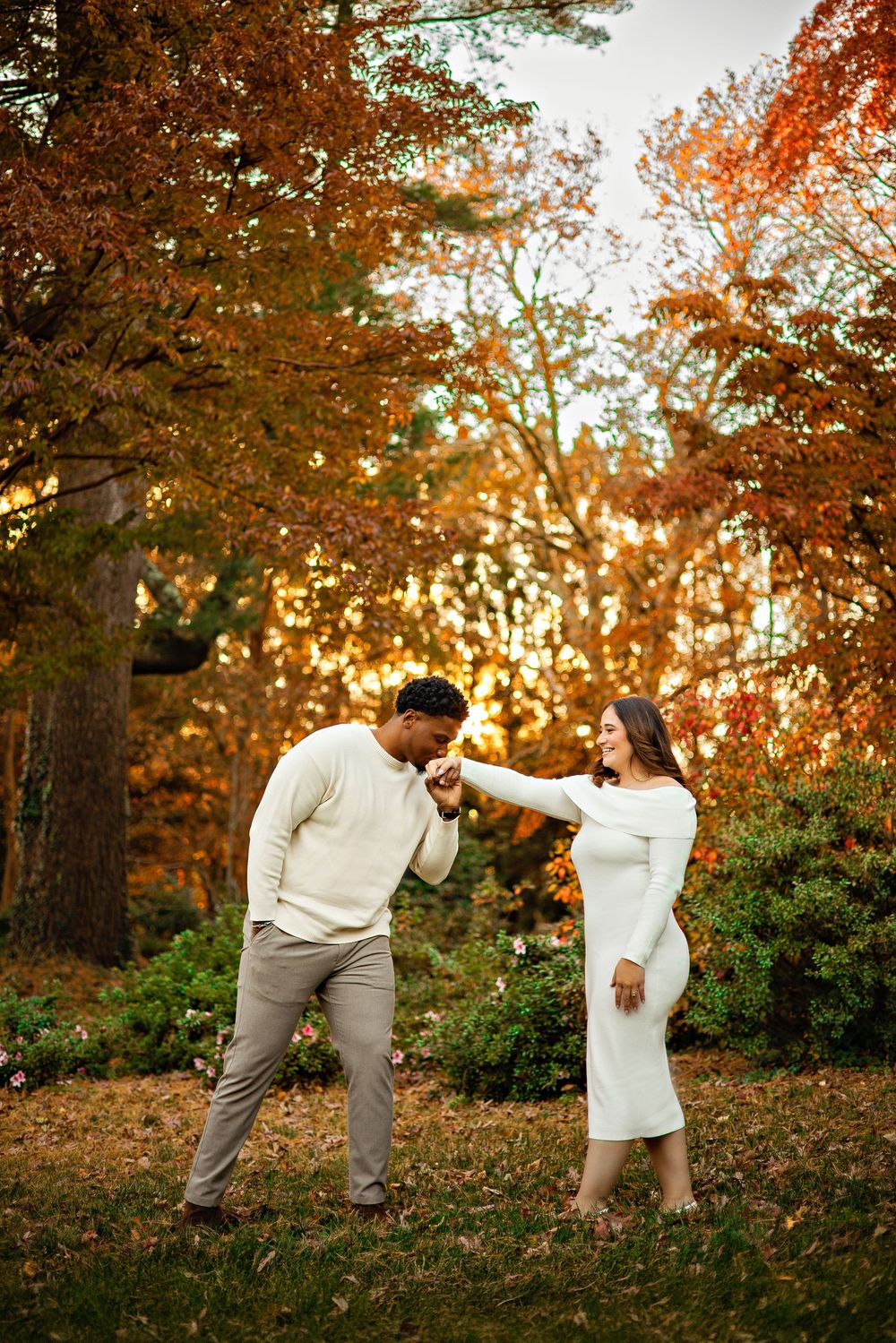 A couple in white and gray outfits playfully poses together in a sunlit autumn forest setting during a photoshoot.