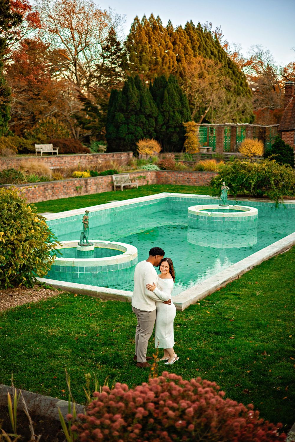 Outdoor swimming pool surrounded by autumn foliage with stone walls and manicured lawn in a garden setting.