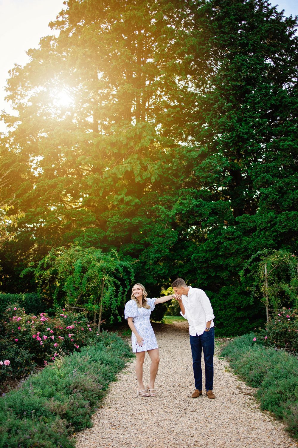 A couple walks along a sunlit gravel path through a garden with evergreen trees and flowering shrubs at sunset.
