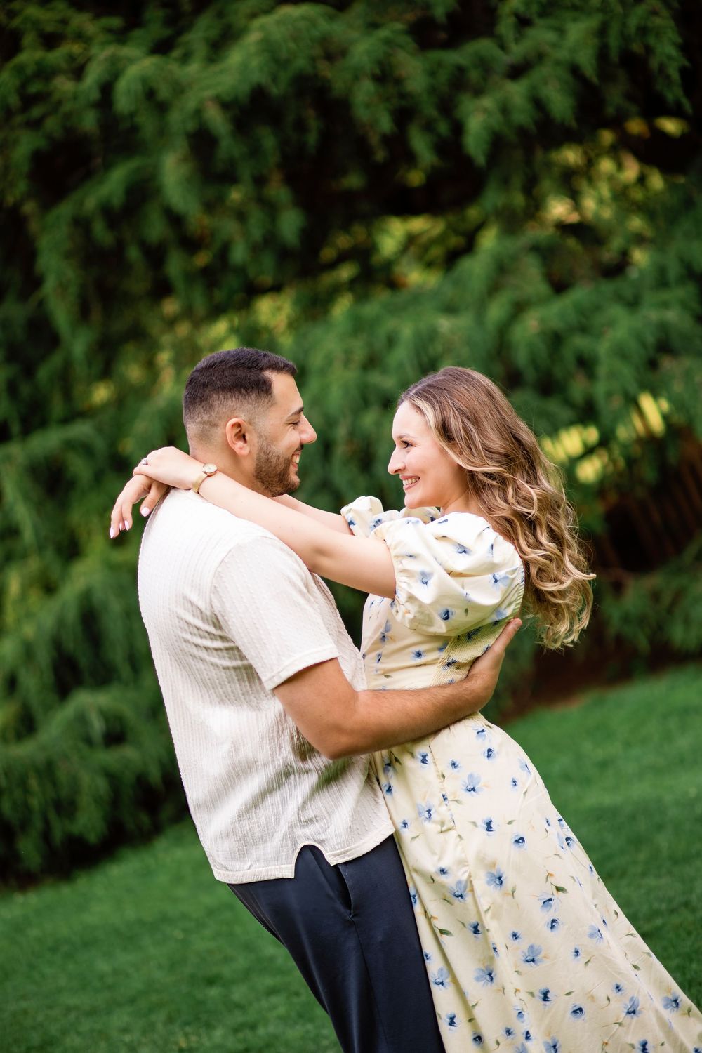 A joyful couple dancing and embracing on a lawn with evergreen trees in the background.