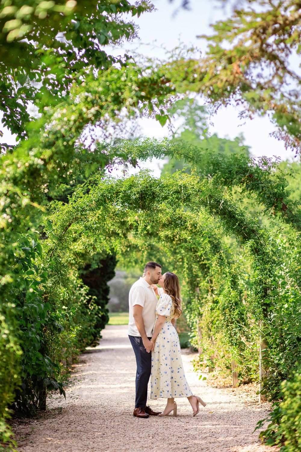 A romantic couple shares a tender moment under a lush green garden archway on a gravel path.