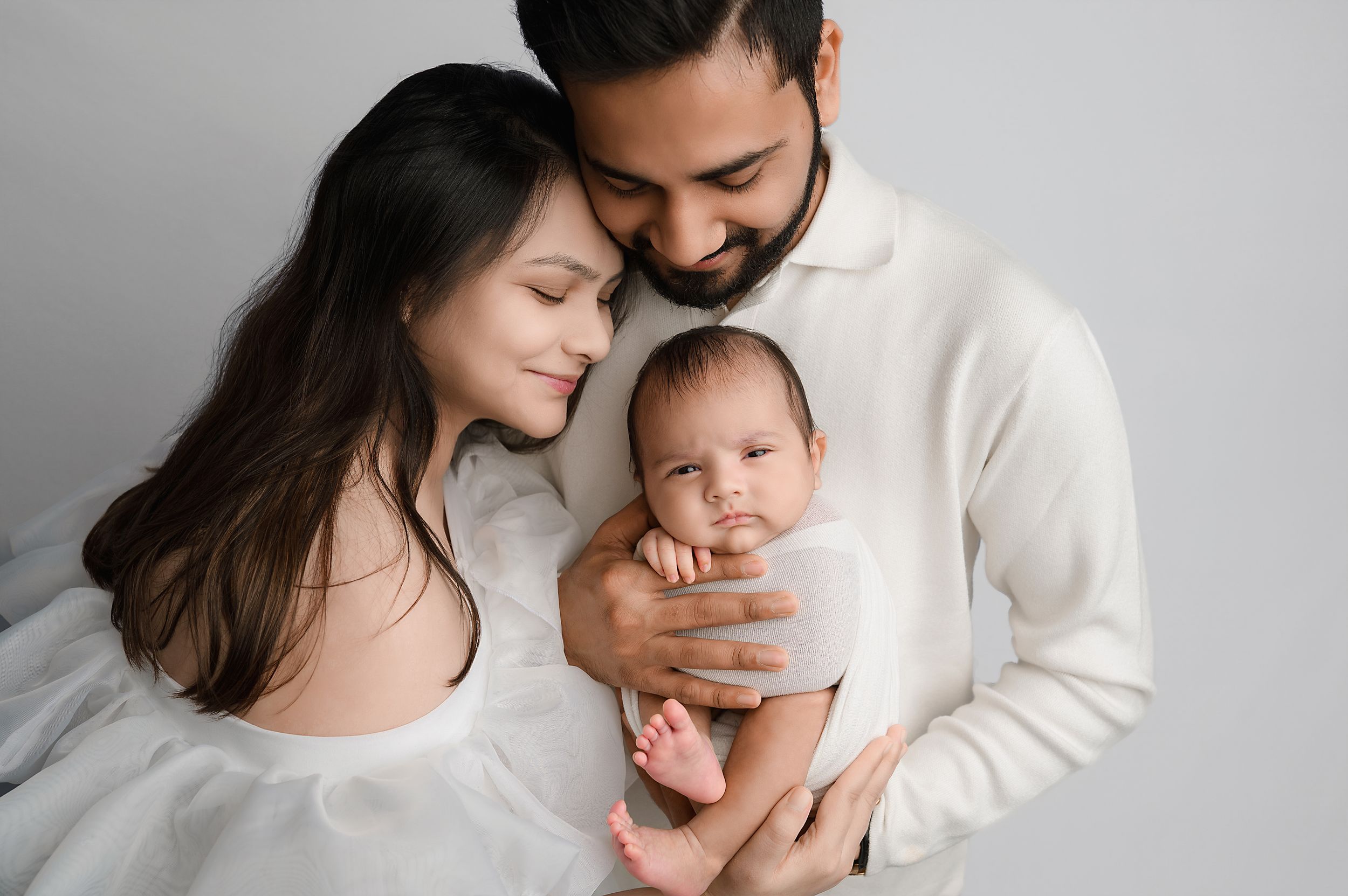 A tender family moment captured in black and white as parents embrace their baby in a minimalist studio setting.