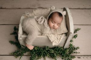A sleeping newborn lies on a white wooden surface surrounded by delicate green ivy leaves and white fabric.