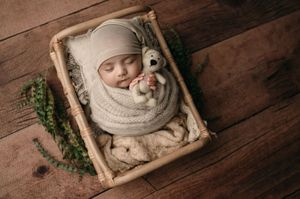 A sleeping infant wrapped in a cozy white knit blanket lies in a rustic wooden basket with greenery beside it.
