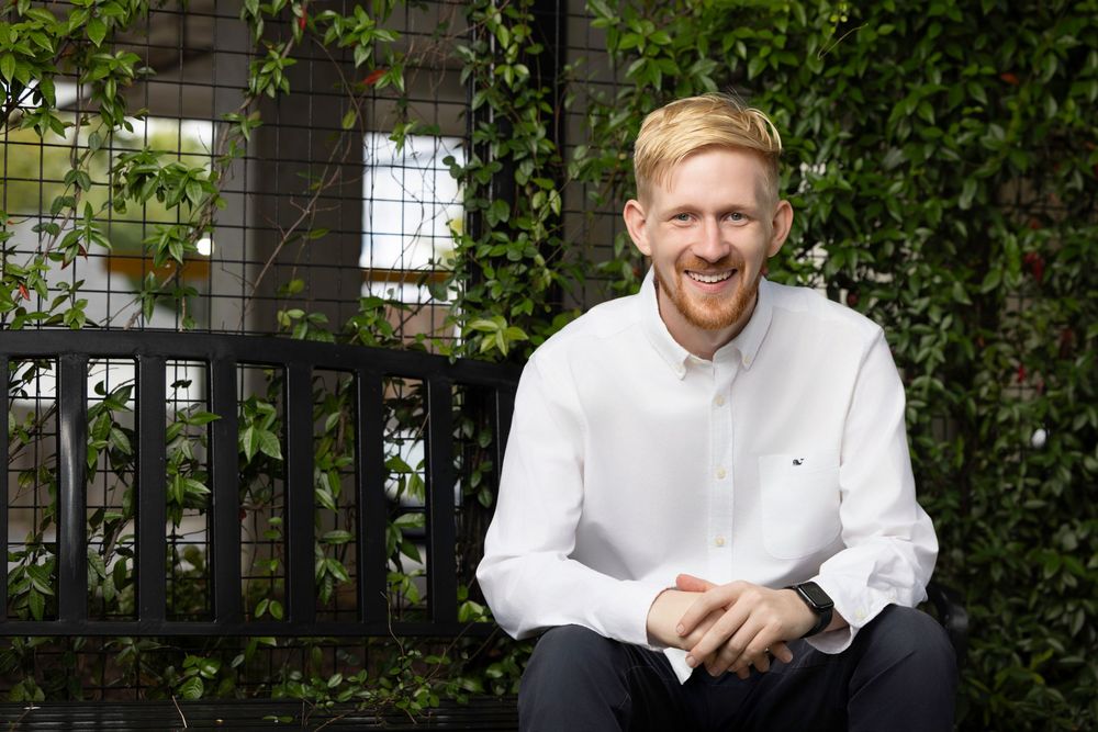A person in a white button-down shirt sits outdoors smiling against a leafy green garden backdrop.