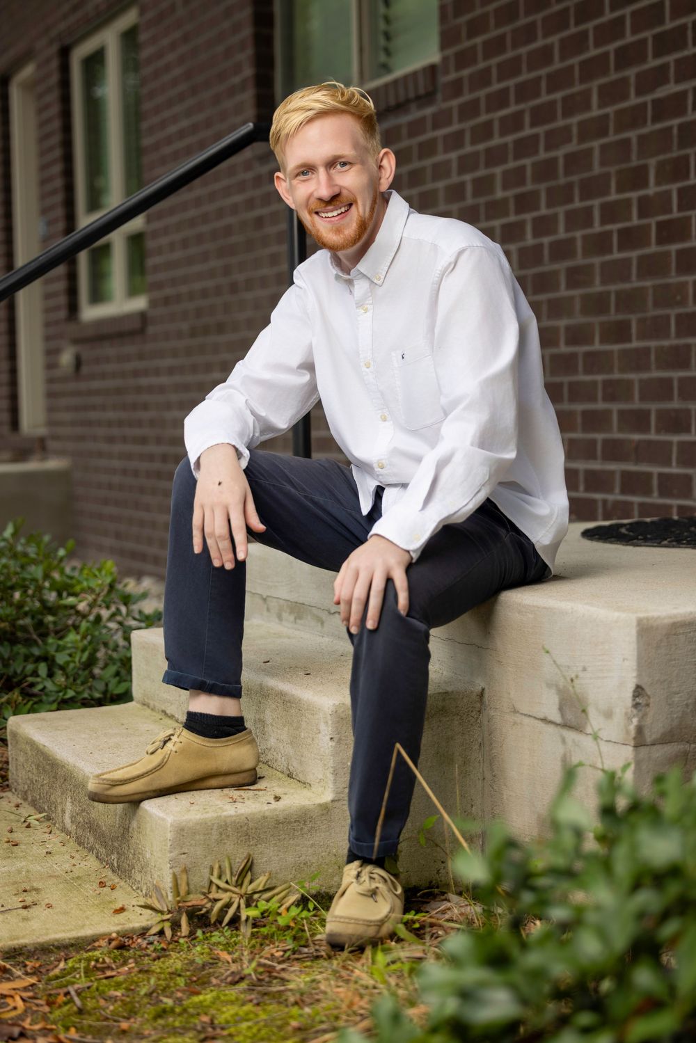 A person sits casually on concrete steps wearing a white button-down shirt and navy pants against a brick building backdrop.