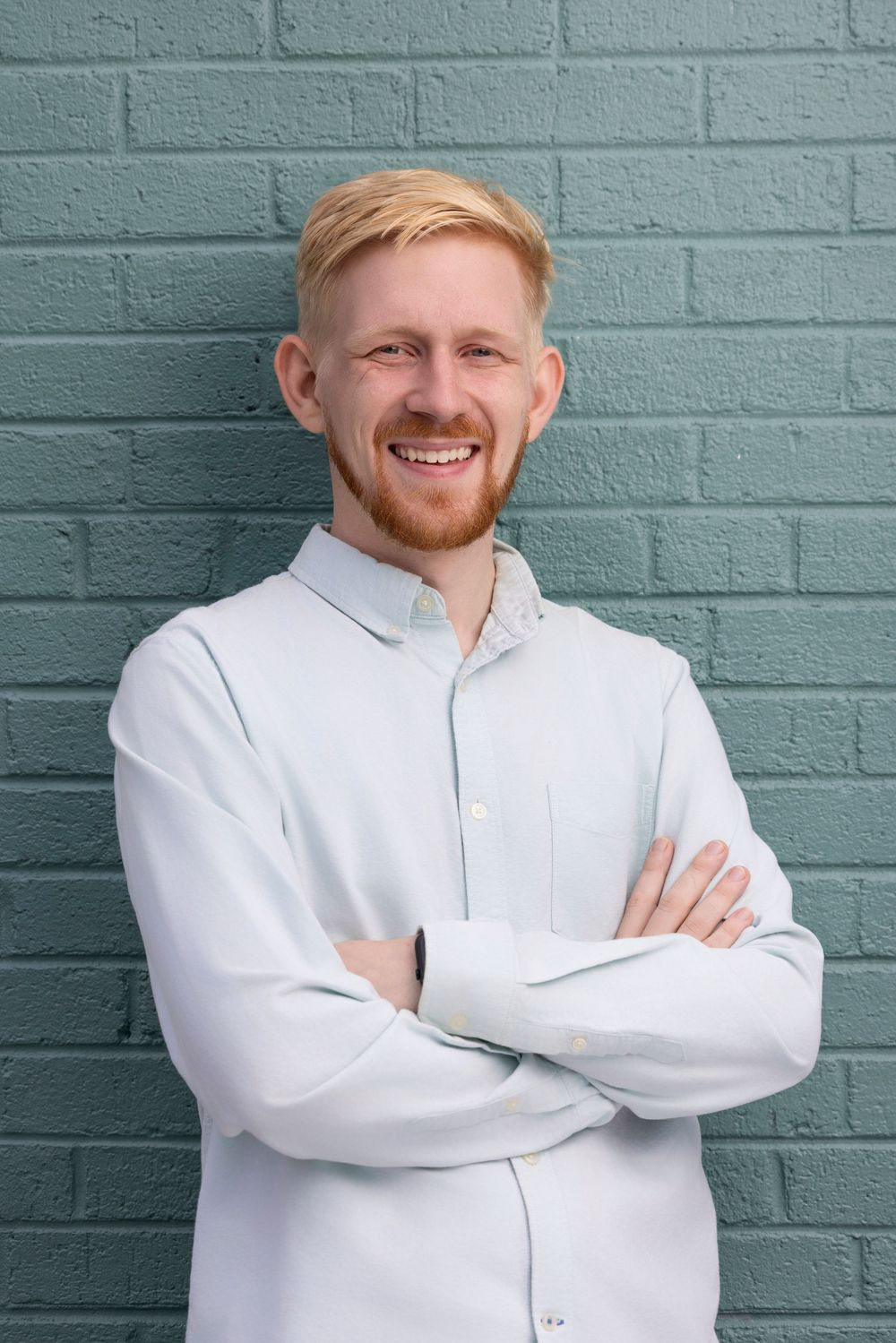 A person in a white button-up shirt poses against a teal painted brick wall with crossed arms and a friendly expression.