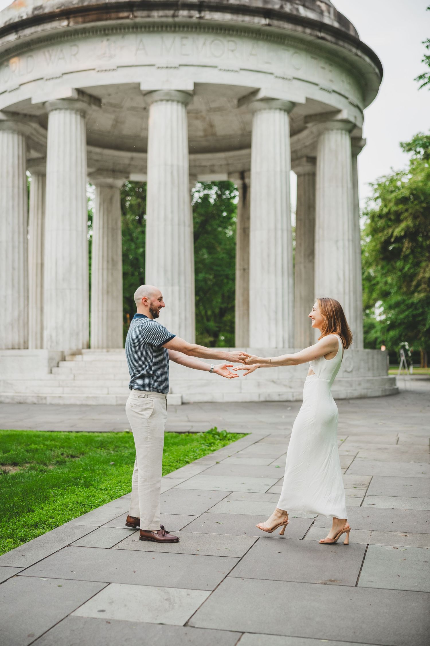 A couple holds hands and spins playfully in front of a neoclassical columned monument on a stone patio.