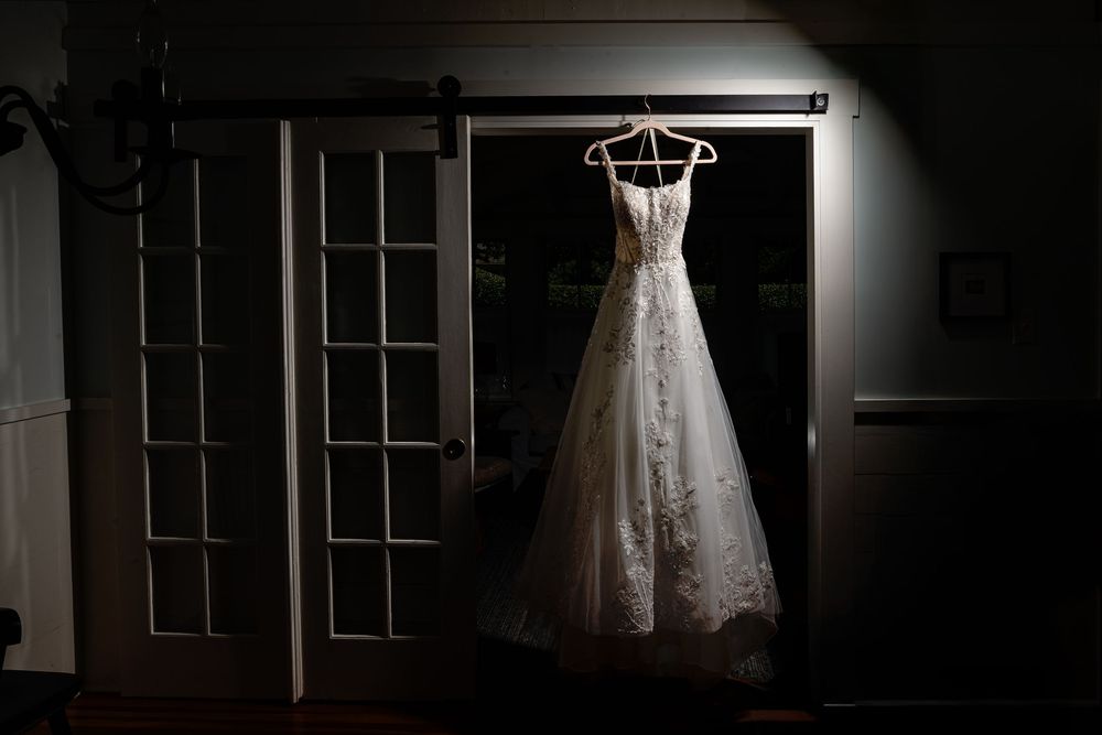 A white lace wedding dress hangs elegantly against a dark wall with window panes casting dramatic shadows.