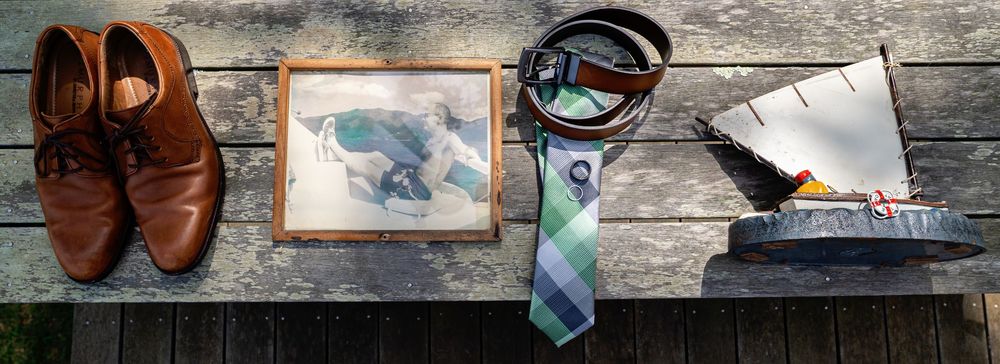 Brown leather dress shoes, blue tie, sunglasses and framed artwork arranged on rustic wooden surface.
