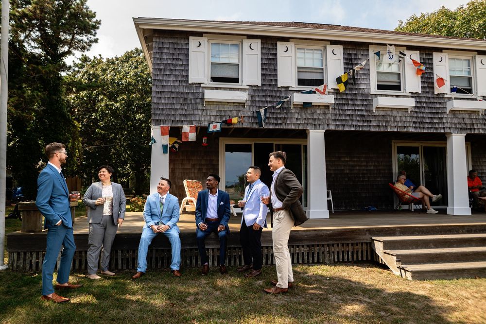 Guests gather outside a gray-shingled building for an outdoor social event on a sunny summer day.