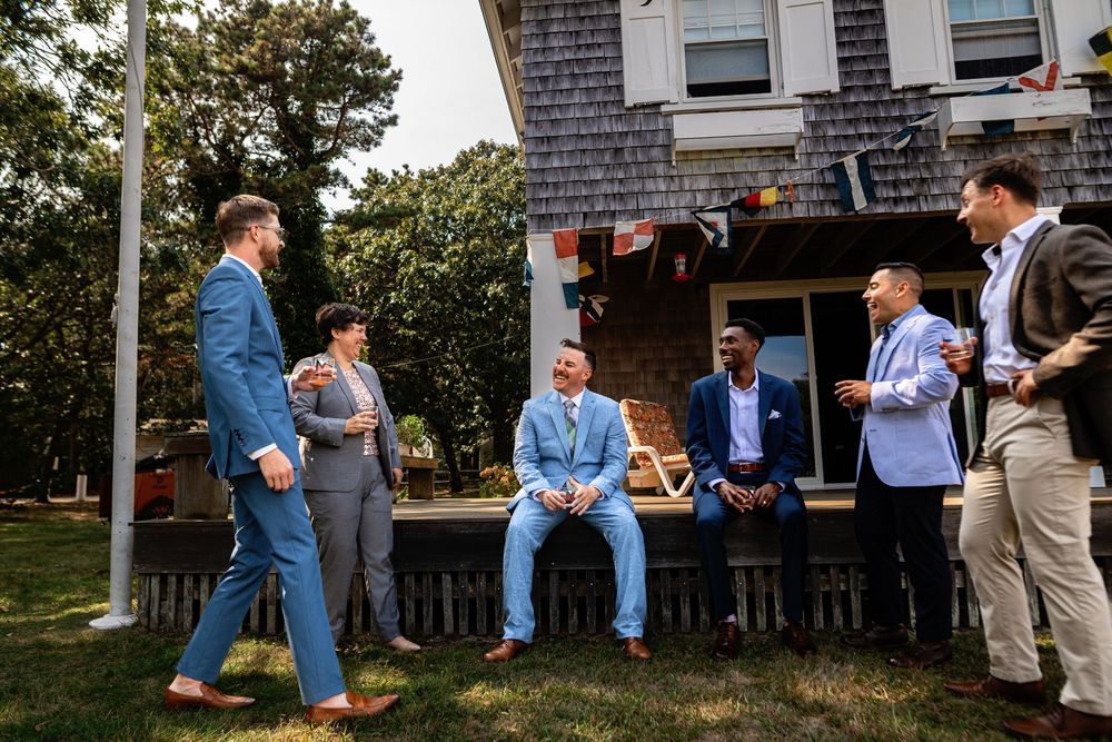 A group gathers outdoors on a sunny day near a wooden house with shingle siding for a casual social event.