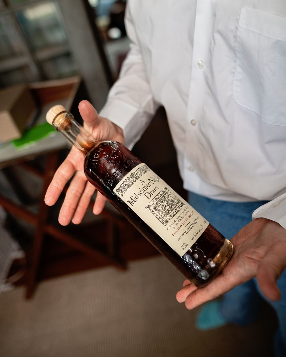A hand holds an aged bottle of wine or spirit against a blurred background.
