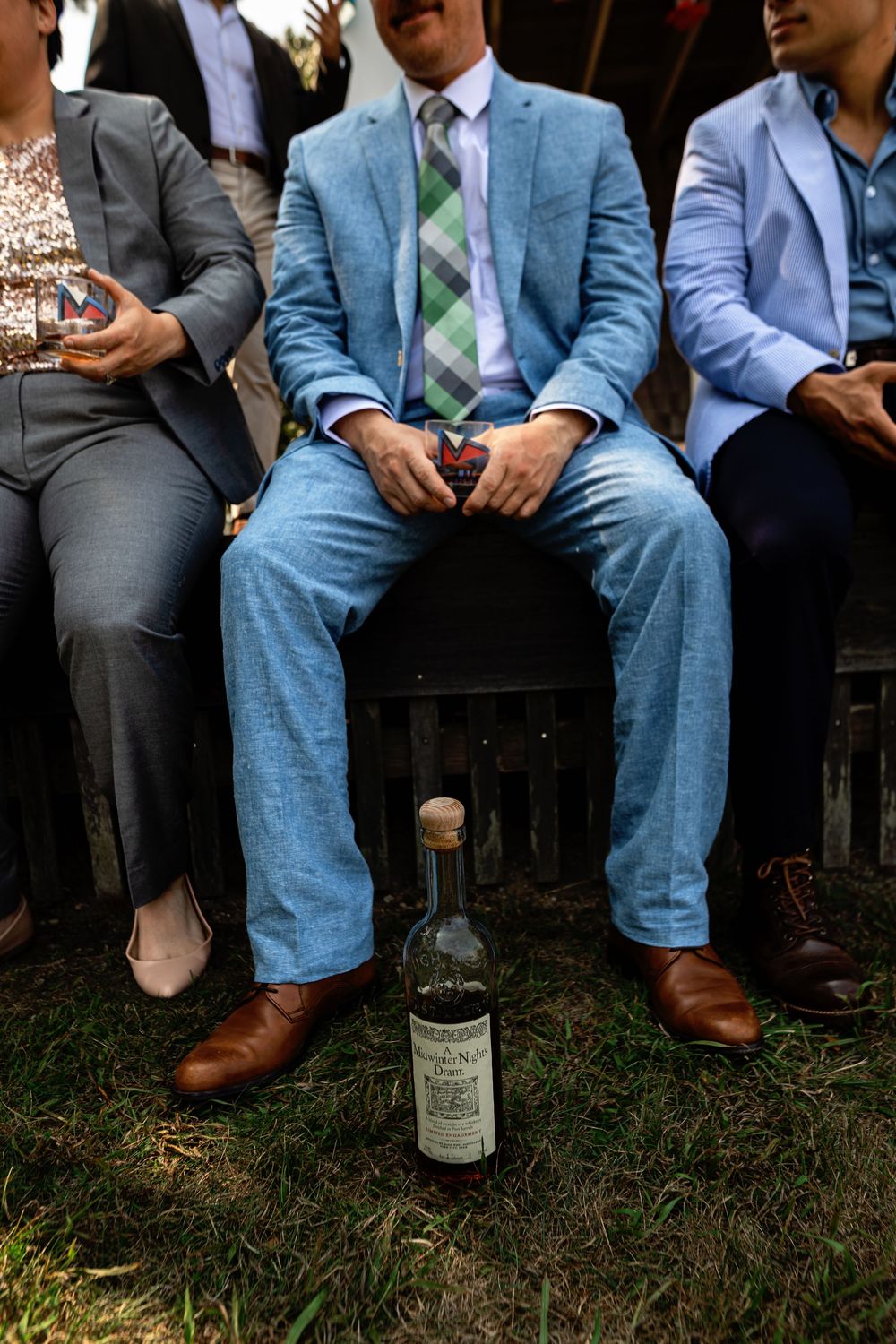 Formal dress shoes and suit pants of wedding party members sitting on grass during outdoor event.