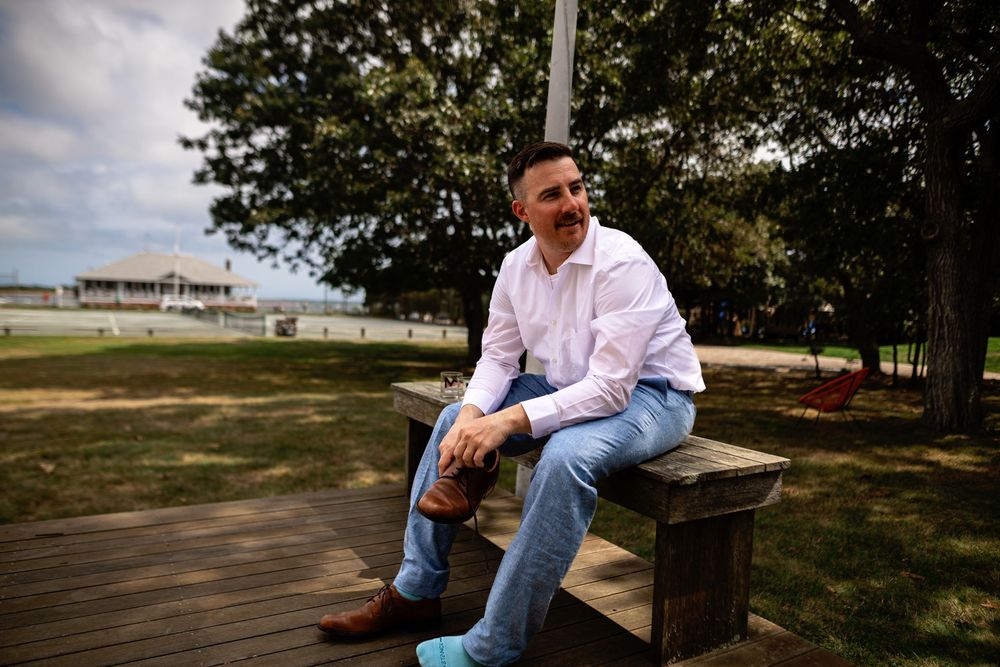 Person in blue jeans and white shirt sits thoughtfully on wooden bench in sunny outdoor setting.