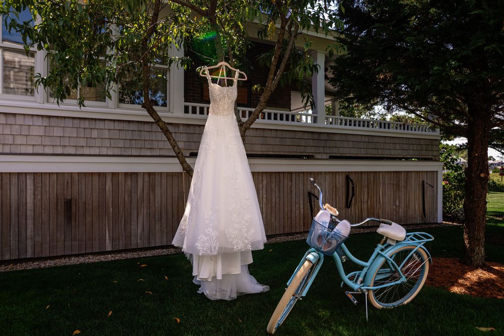 A white wedding dress hangs from a tree branch with a turquoise bicycle and wooden fence in the background.