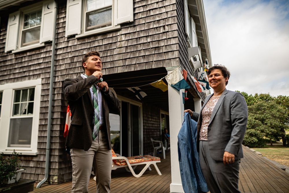 Wedding party members share laughs and candid moments on a gray-shingled porch during preparations.
