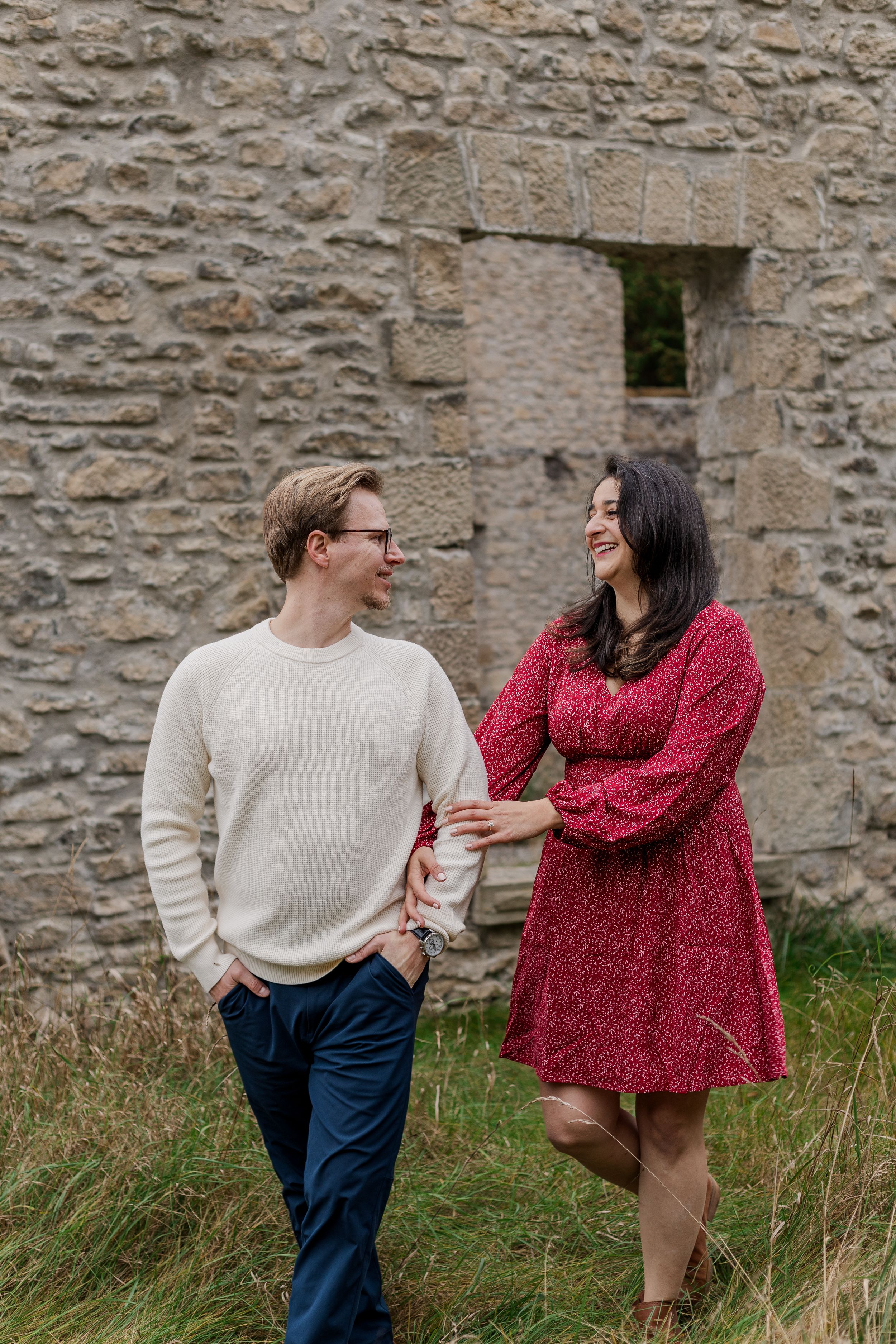 A couple in casual attire shares a playful moment against a rustic stone wall backdrop.