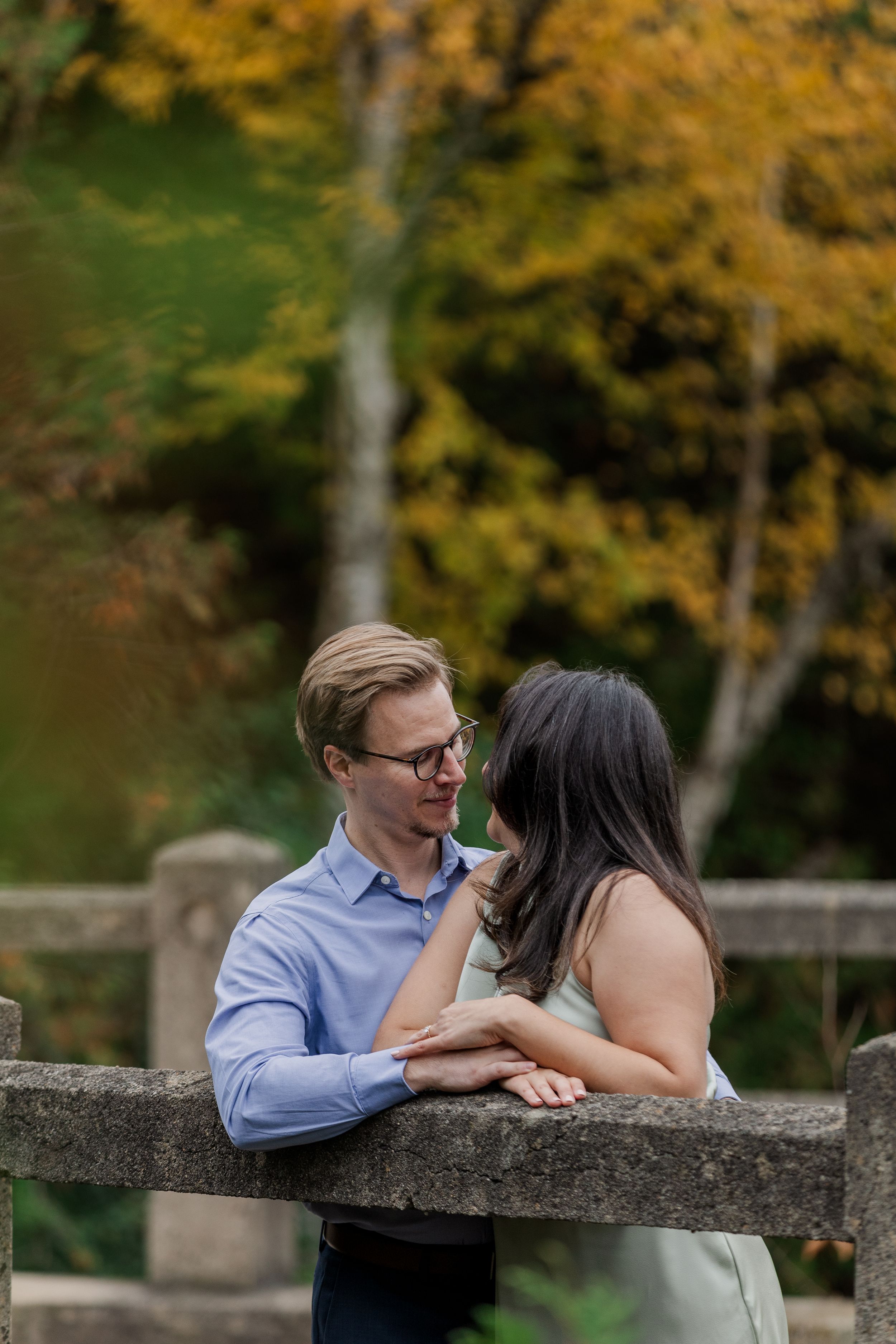 A romantic couple shares tender moments while leaning against a stone wall surrounded by autumn foliage.