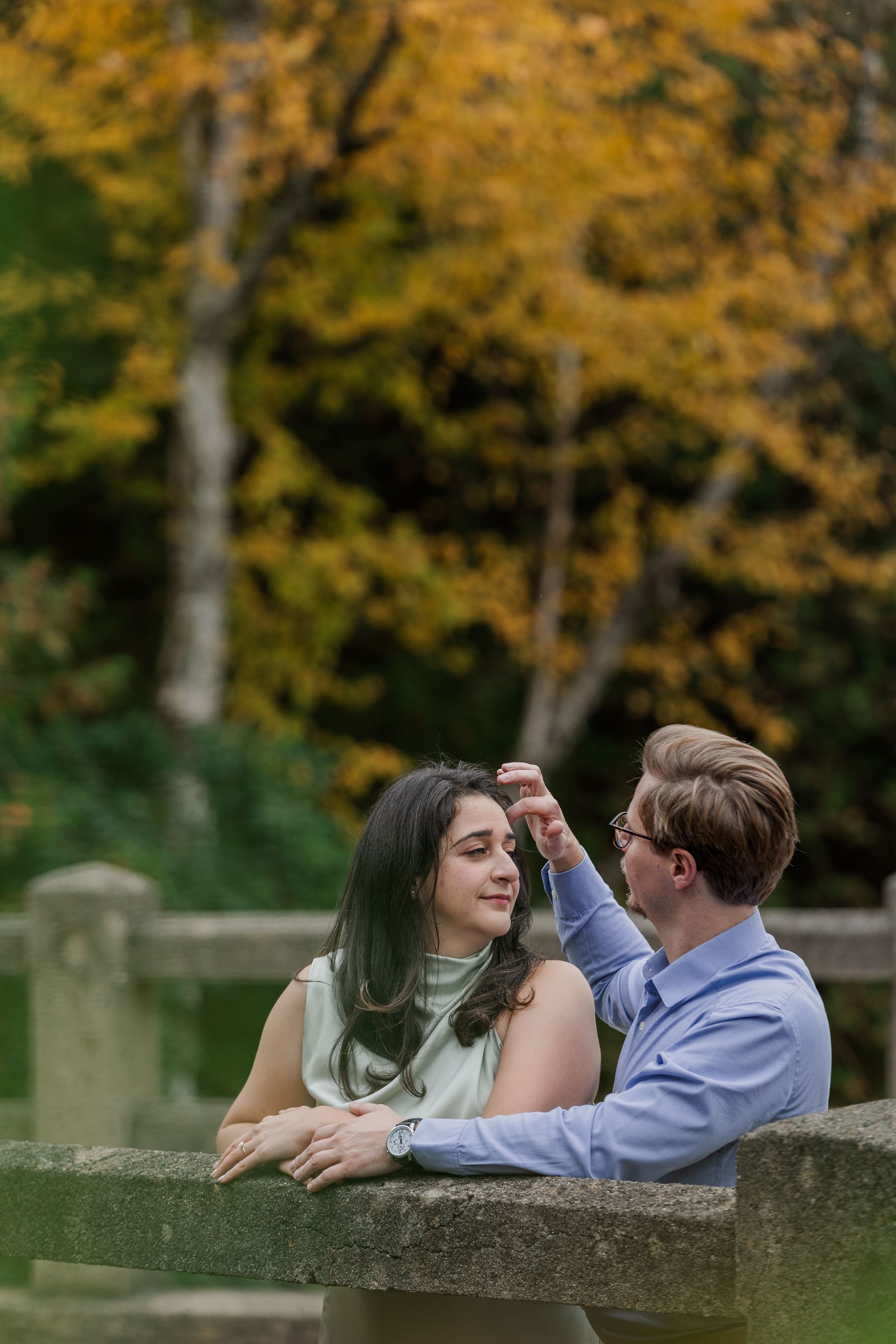 A couple lies together on a stone wall surrounded by autumn trees with golden foliage in the background.