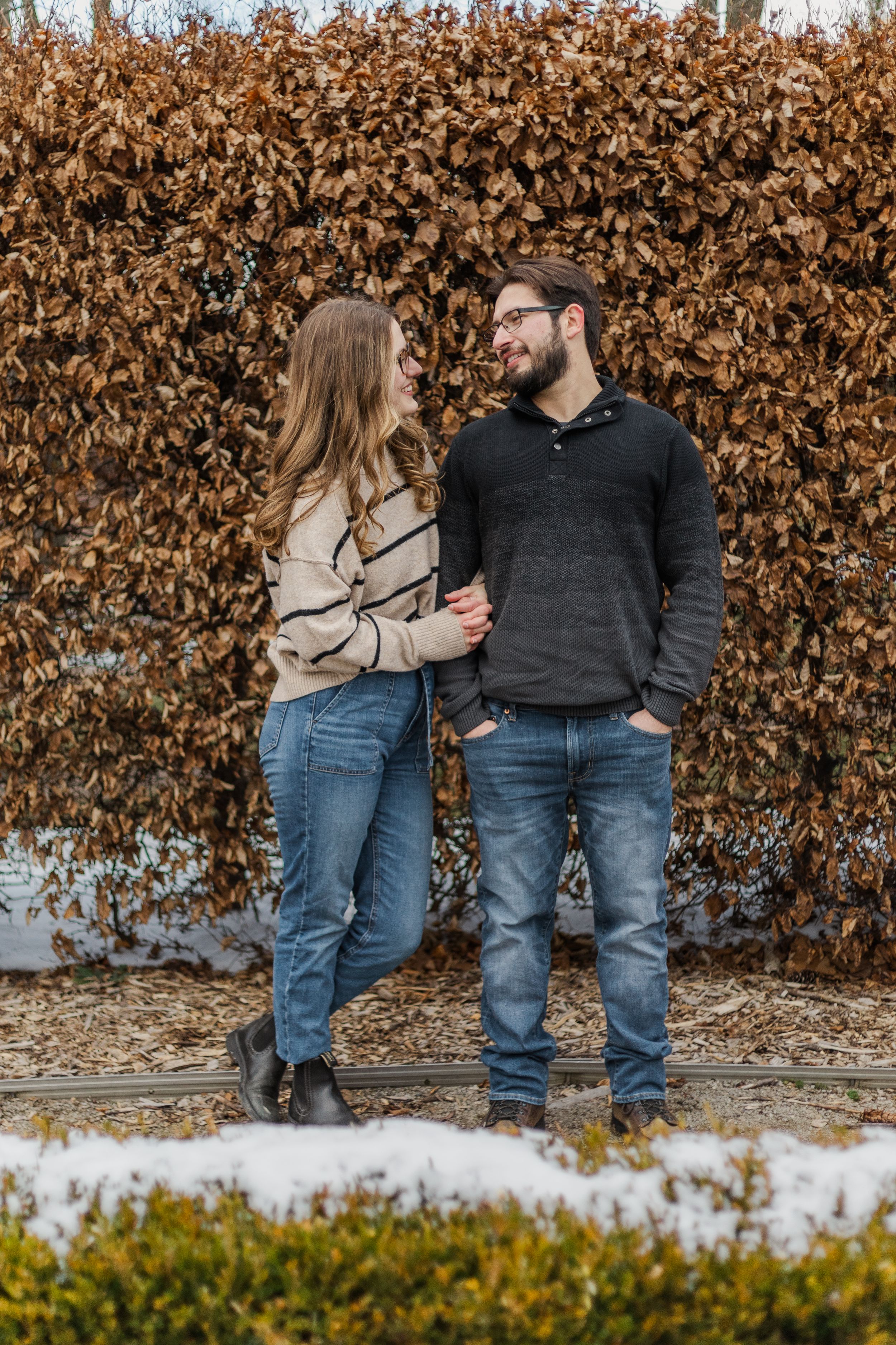 A couple stands together against a brown hedge wall in winter with snow on the ground.